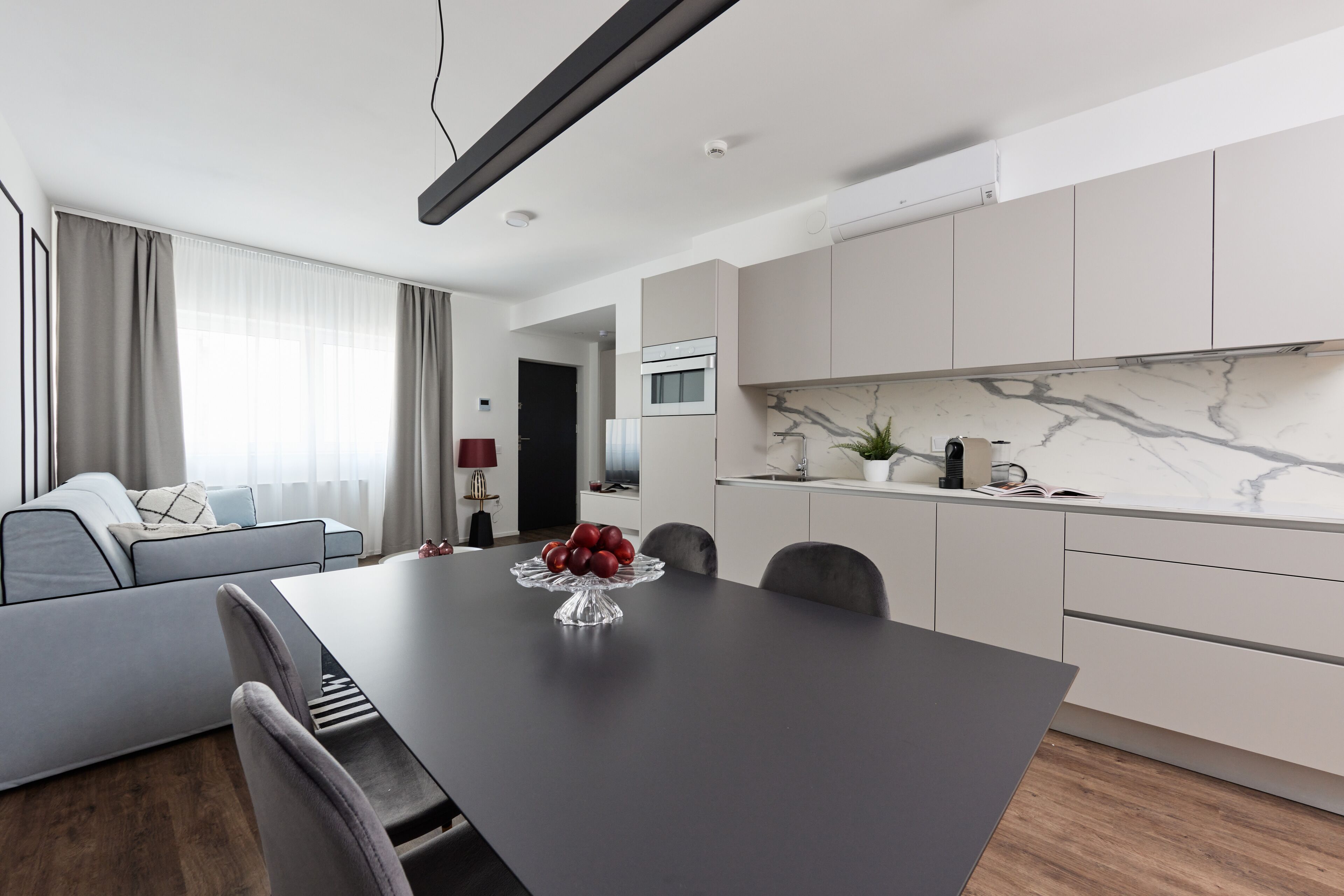 Ein modernes Wohnzimmer mit einem grauen Sofa, einem Esstisch mit Obstschale und einer Küche mit weißen Schränken und einer Marmor-Spritzschutzwand.