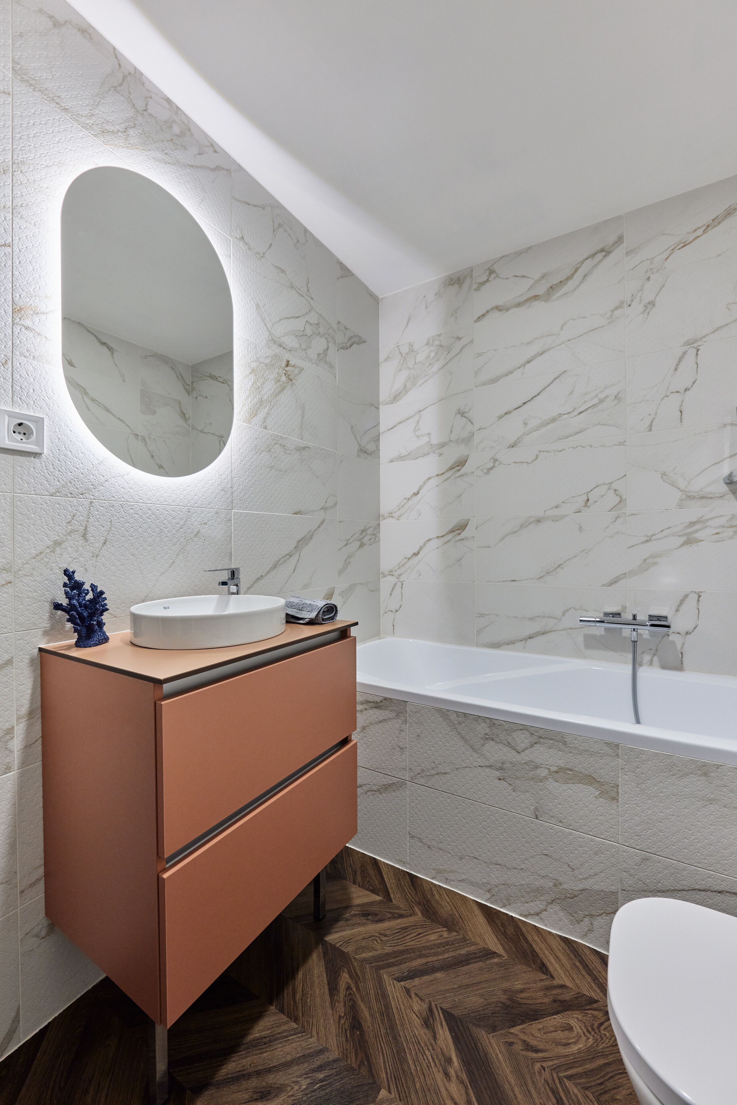 A modern bathroom with a marble wall, a round mirror, a wooden vanity, a bathtub, and a herringbone-patterned floor.