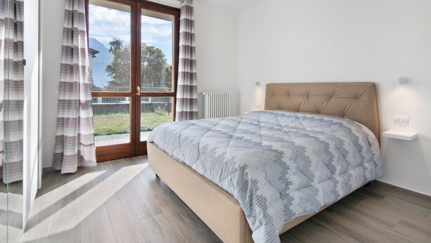 Ein Schlafzimmer mit Bett, einem Fenster mit Vorhängen, einem Heizkörper und Blick auf Grünflächen und Berge.