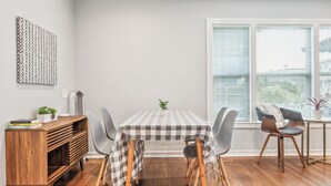Un coin repas avec une nappe à carreaux, des chaises modernes, un buffet en bois et une fenêtre avec des stores.