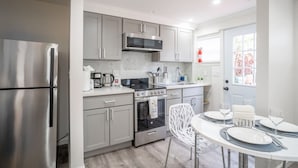 A modern kitchen with stainless steel appliances, white cabinetry, and a dining area with a white table set for two. - Luxo Stays |Modern Studio| Near SFO, Westlake BART (Daly City)