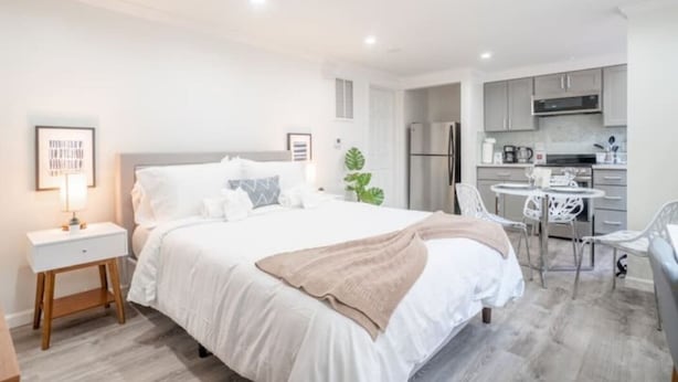 A modern hotel room with a neatly made bed, a small bedside table, a chair, and a kitchenette area. - Luxo Stays |Modern Studio| Near SFO, Westlake BART (Daly City)
