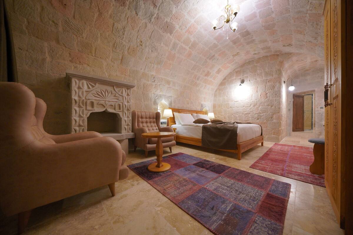 A cozy room with a stone wall, a bed, a chair, a small table, and a fireplace.