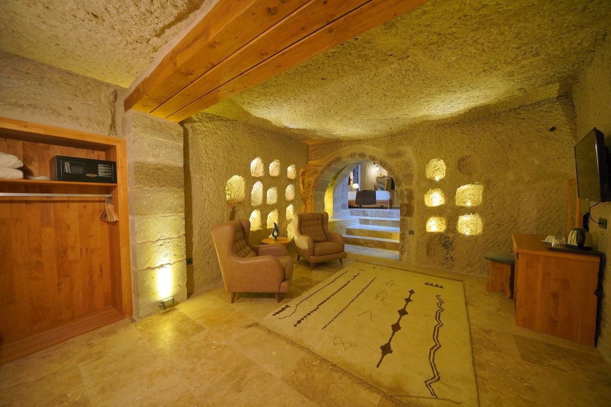 A stone-walled room with two leather chairs, a wooden bench, a patterned rug, and a wooden cabinet with a television.