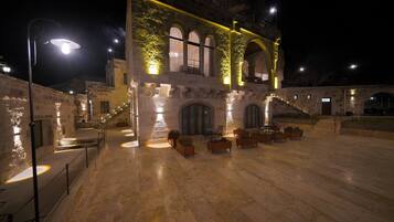 An outdoor courtyard with stone flooring, seating areas, and a building with arched windows and a staircase.