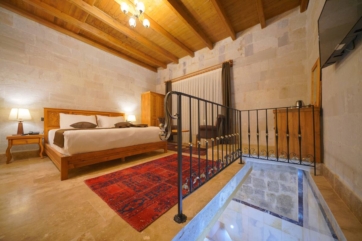 A bedroom with a wooden bed, a red rug, a staircase with a metal railing, and stone walls.