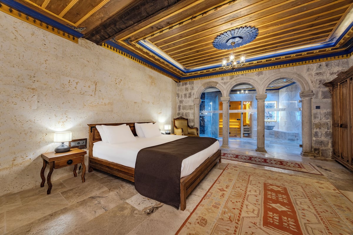 A bedroom with a large bed, wooden headboard, and a nightstand with a lamp. There is a patterned rug on the floor and a view of a courtyard with columns and a fountain.