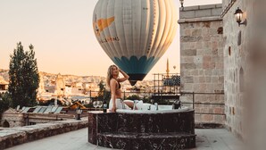 Un globo aerostático con la inscripción "Guerin Balloons", una mujer sentada sobre una plataforma de mármol y un paisaje urbano al fondo.