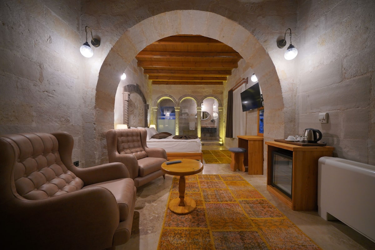 A cozy room with a sofa, armchair, and a small table. There is a bed with a TV in the background.