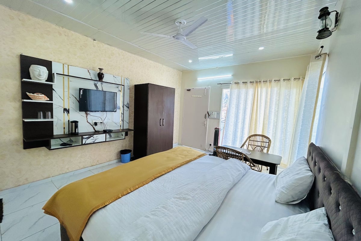 A hotel room with a bed, a TV mounted on the wall, a wardrobe, a table with chairs, and a window with curtains.