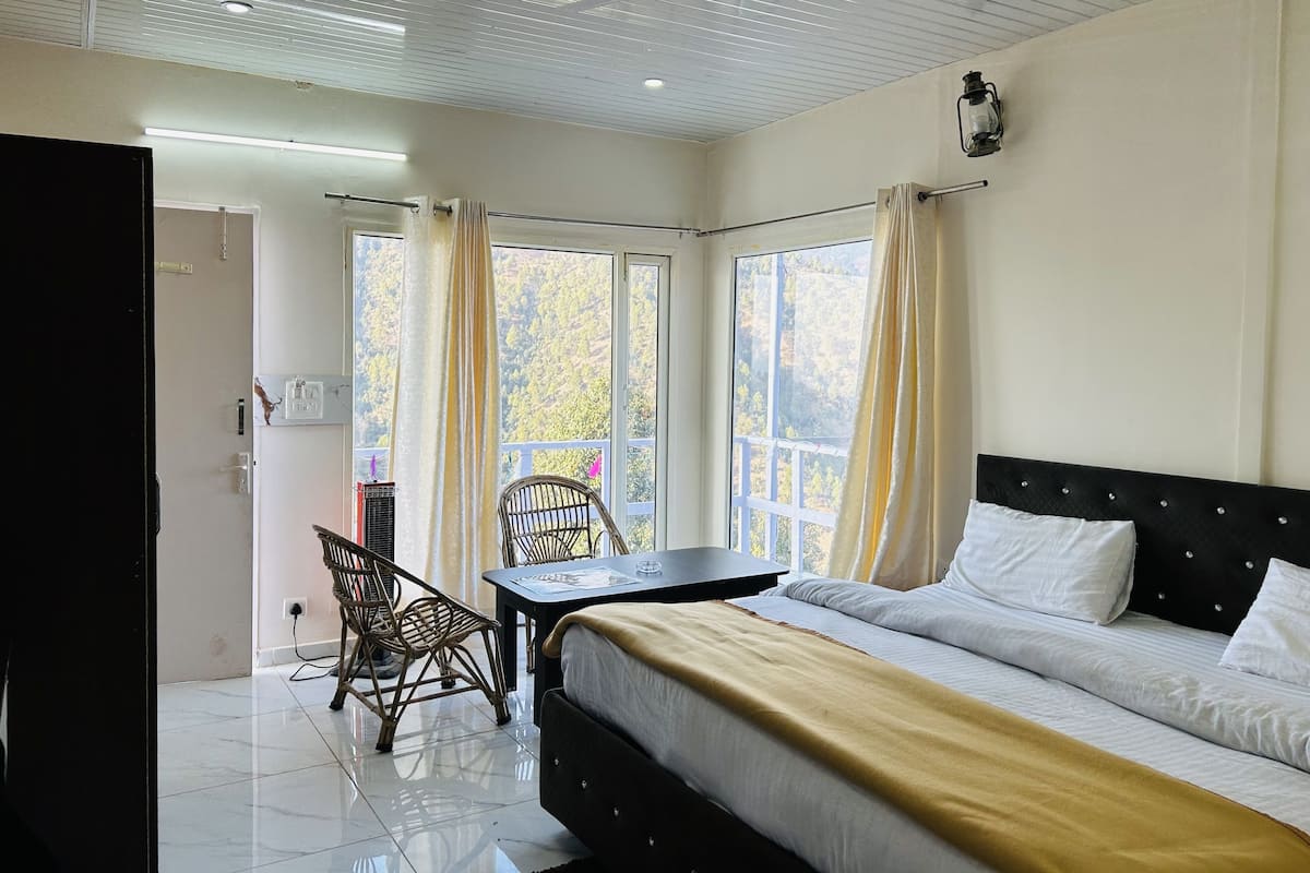 A hotel room with a bed, a table, a chair, a ceiling fan, and a view of trees through the windows.