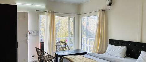 A hotel room with a bed, a table, and a chair. There is a window with curtains, a ceiling fan, and a light fixture.