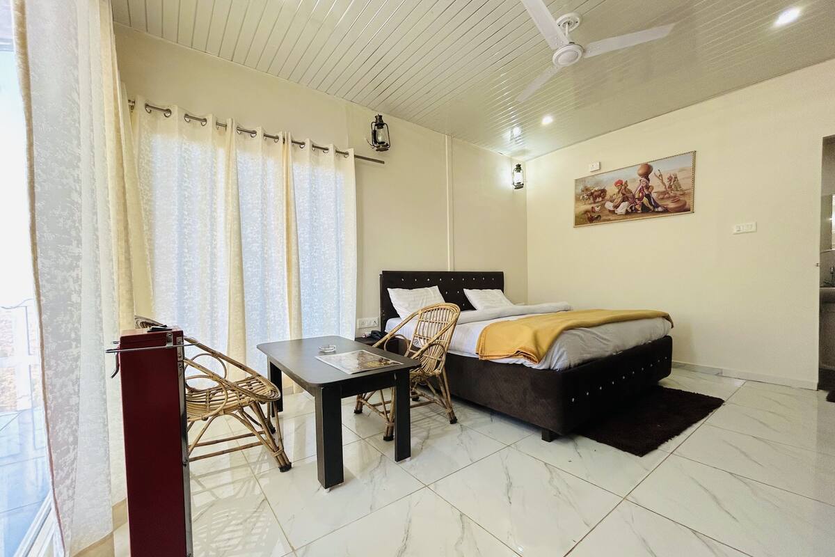 A bedroom with a bed, a table, a chair, a window with curtains, a ceiling fan, and a painting on the wall.