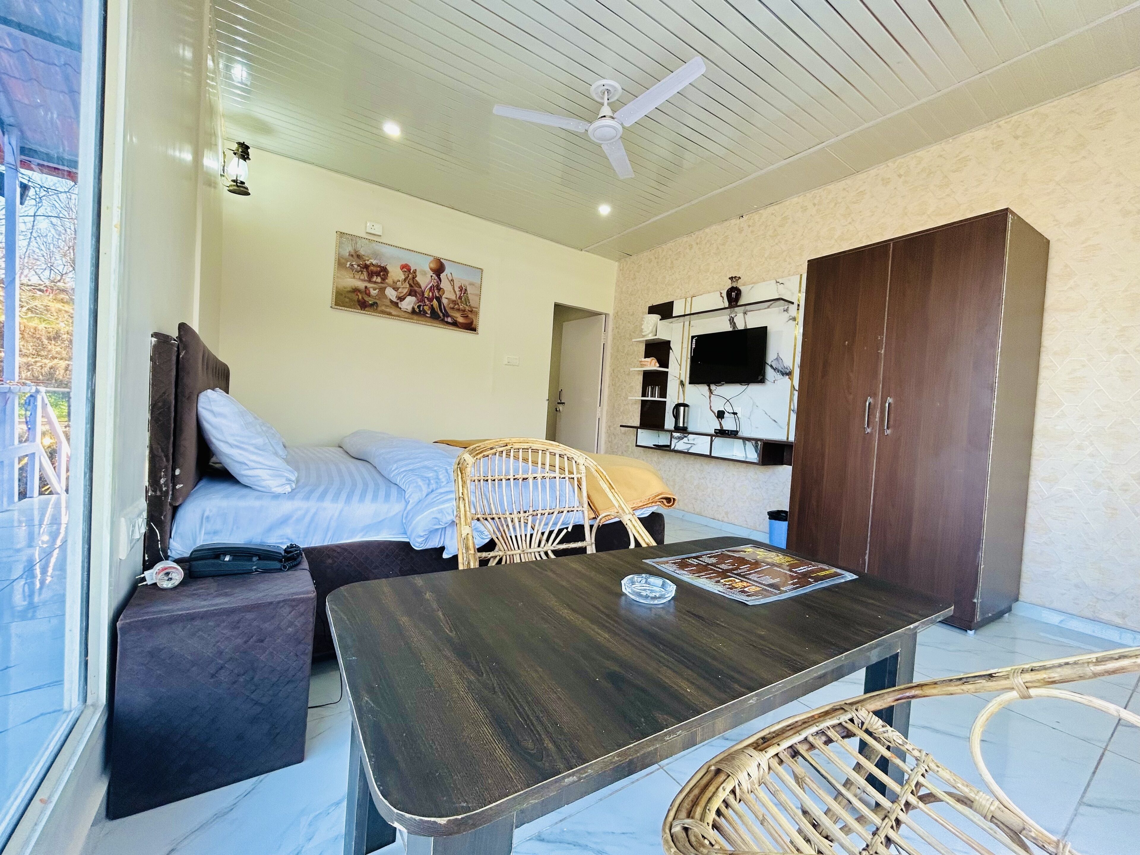 Un dormitorio con cama, una mesa de madera, una silla de mimbre, un armario y un televisor montado en la pared, además de un cuadro.