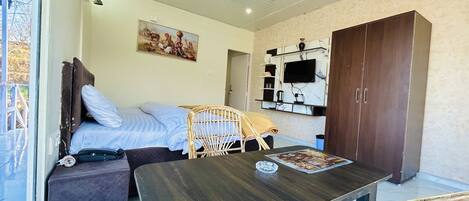 Un dormitorio con cama, una mesa de madera, una silla de mimbre, un armario y un televisor montado en la pared, además de un cuadro.