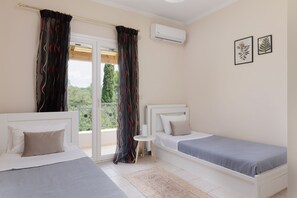 A bedroom with two beds, a small bedside table, a window with curtains, and an air conditioning unit. - Country Villa Verde (Corfu)