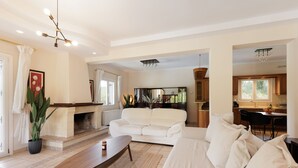 A spacious living room with a white sofa, a wooden coffee table, and a fireplace. There is a kitchen visible in the background with wooden cabinets and a dining area. - Country Villa Verde (Corfu)