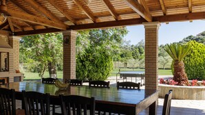 An outdoor patio with a wooden table and chairs, surrounded by brick pillars and a view of greenery and a golf course. - Country Villa Verde (Corfu)