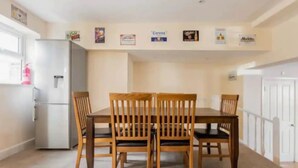 A dining area with a wooden table and chairs, a refrigerator, and a white door. - Abbey House SW London for Big Groups (Colliers Wood)