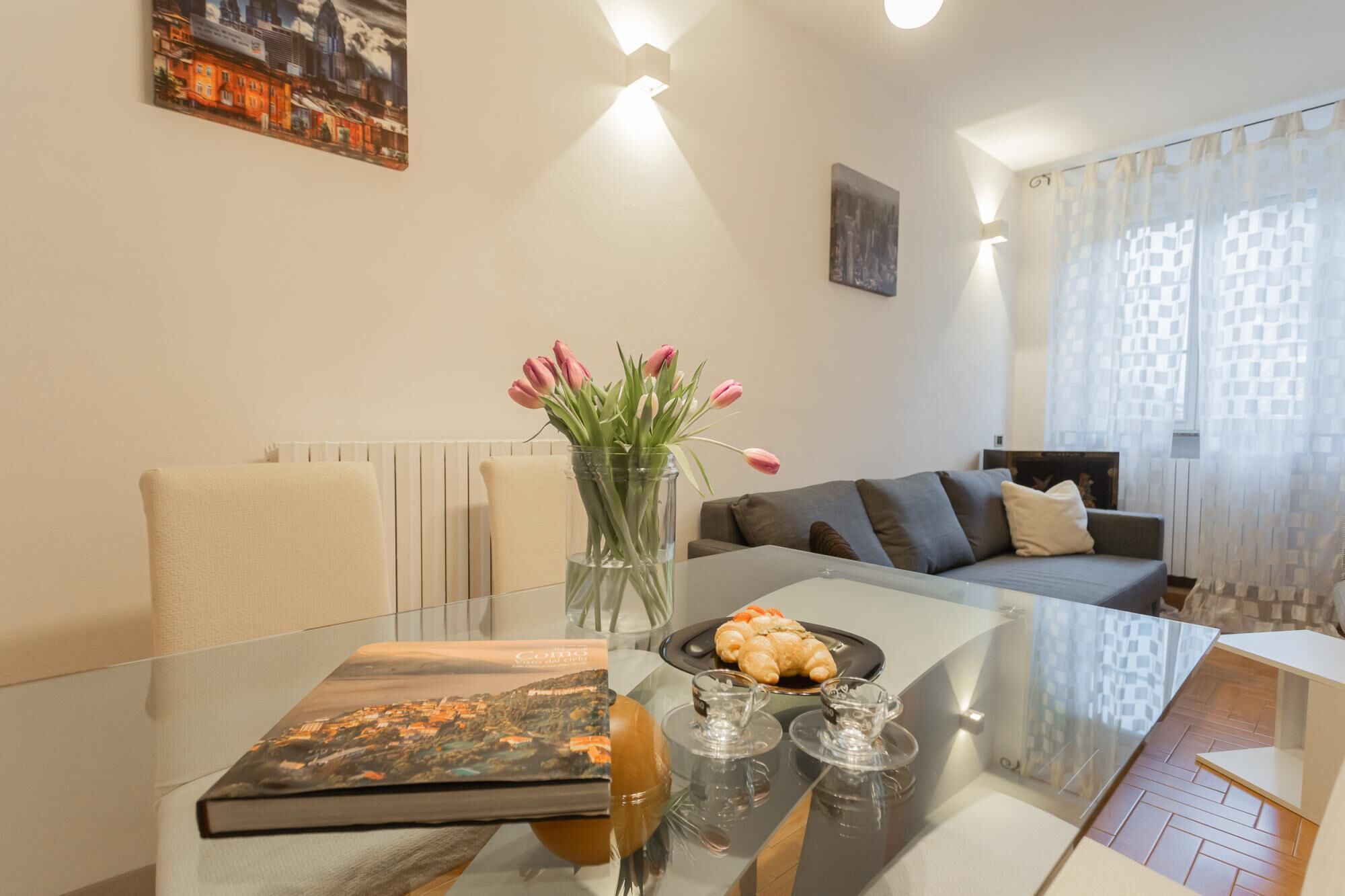 A modern living room with a glass coffee table, a sofa, a vase with tulips, and a framed picture on the wall.