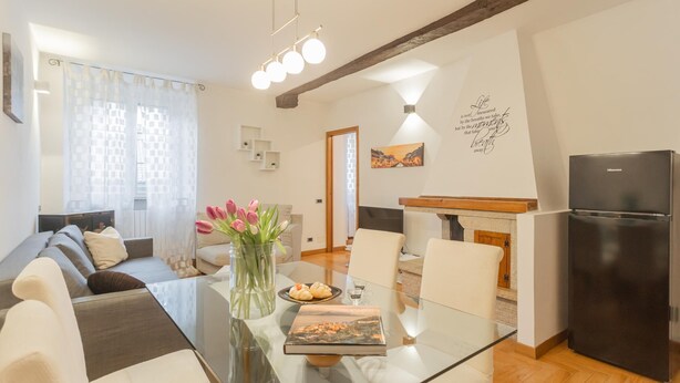 Un espace de vie moderne comprenant une table à manger en verre, un canapé, un coin cuisine avec un réfrigérateur et une vue sur la ville par une fenêtre.