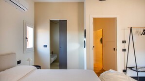 Una habitación de hotel con una cama grande, una mesita de noche, un equipo de aire acondicionado, un armario y un baño visible a través de una puerta abierta.