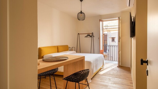 Ein Schlafzimmer mit einem Bett, einem Holztisch, zwei Stühlen, einer Pendelleuchte und Blick ins Freie.