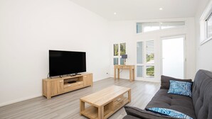 Een moderne woonkamer met een flatscreen televisie op een houten standaard, een grijze bank met gekleurde kussens en een houten salontafel.
