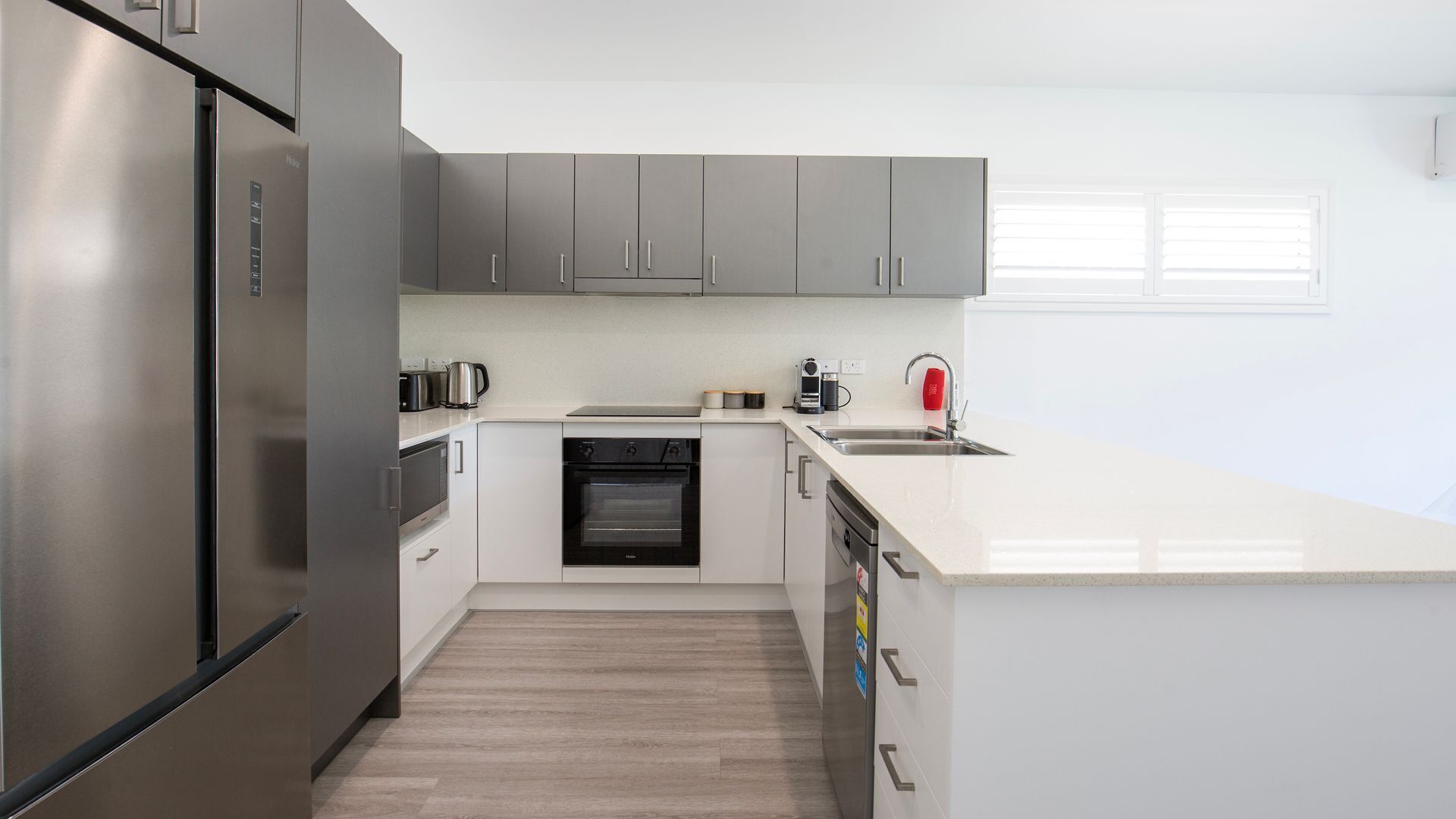 A modern kitchen with stainless steel appliances, grey cabinets, a white countertop, and a window with blinds.