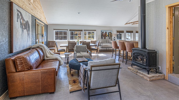 A spacious living room with a leather sofa, a blue ottoman, and a black wood-burning stove. There are multiple chairs and a dining table in the background.