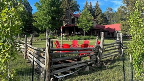 A rustic lodge with a stone exterior and a red roof, surrounded by a wooden fence and red chairs. - Rancho Sendero Correo -Historic Home on 300 Acres- Hot Tub/Pool Table/Trails (Hesperus)