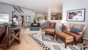 A spacious living room with a brown leather sofa, a wooden coffee table, and a patterned rug. There is a bookshelf filled with books and a chandelier hanging from the ceiling. - Rancho Sendero Correo -Historic Home on 300 Acres- Hot Tub/Pool Table/Trails (Hesperus)