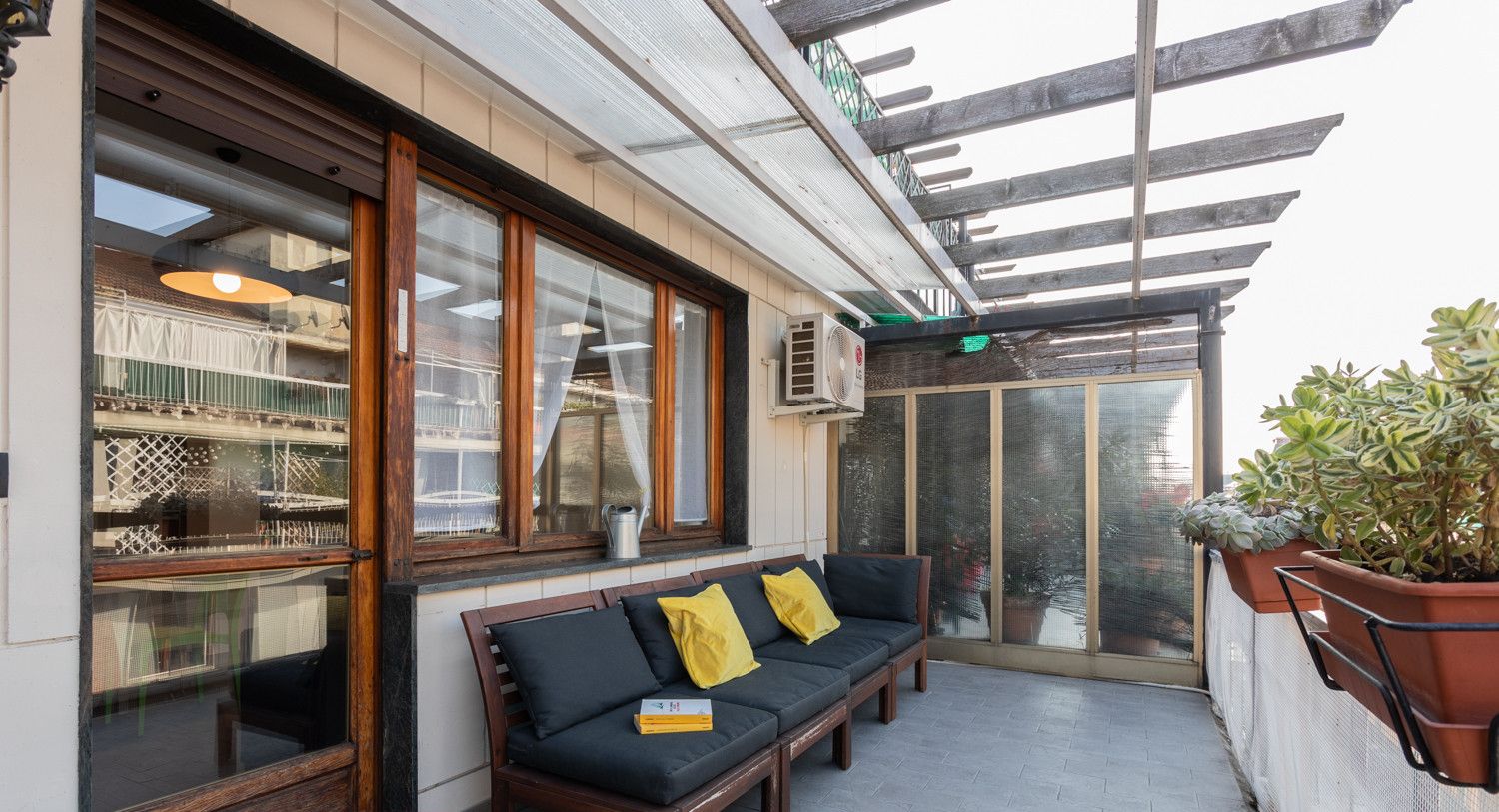 Een overdekt terras met een bank en kussens, een potplant en uitzicht op de straat.