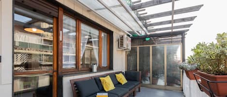 A covered outdoor seating area with a bench and cushions, a potted plant, and a view of the street.