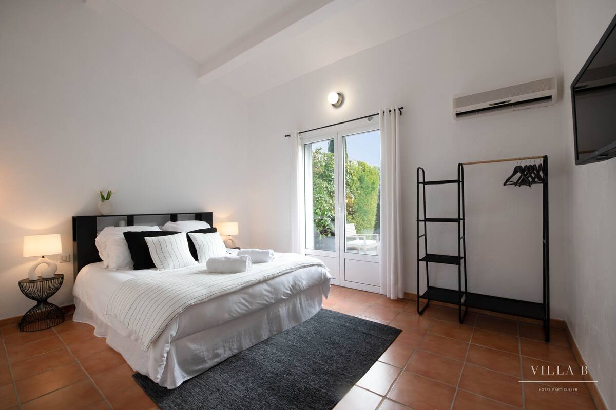 Una camera da letto con un letto, un comodino, una lampada, una finestra con tende, un condizionatore d'aria a parete e una rastrelliera in metallo nero.