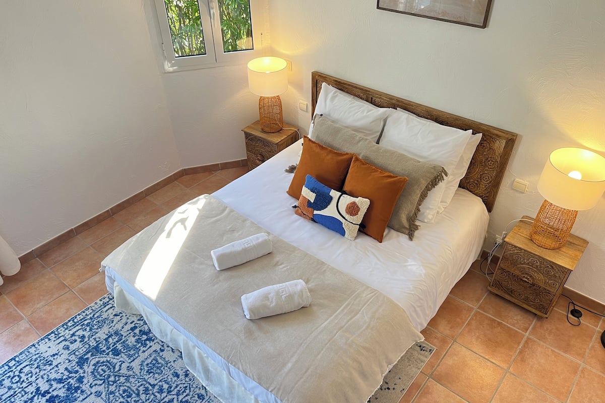 Un letto rifatto con lenzuola bianche, due abat-jour e un tappeto blu con motivo decorativo.