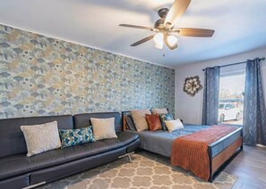 A bedroom with a bed, a sofa, a ceiling fan, a window with curtains, and a patterned wall.