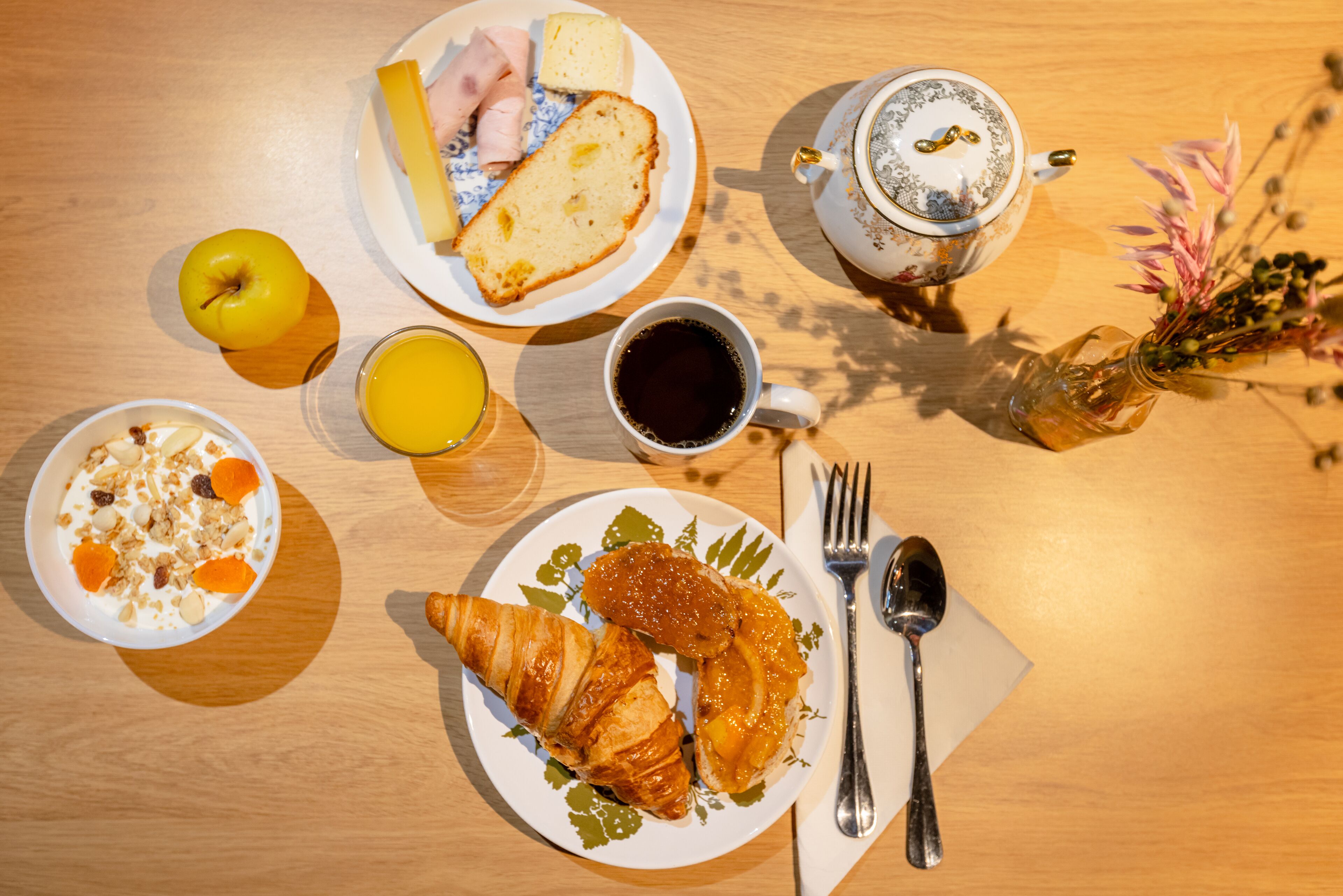 Una mesa de desayuno con croissants, fruta, una taza de café y un florero con flores.
