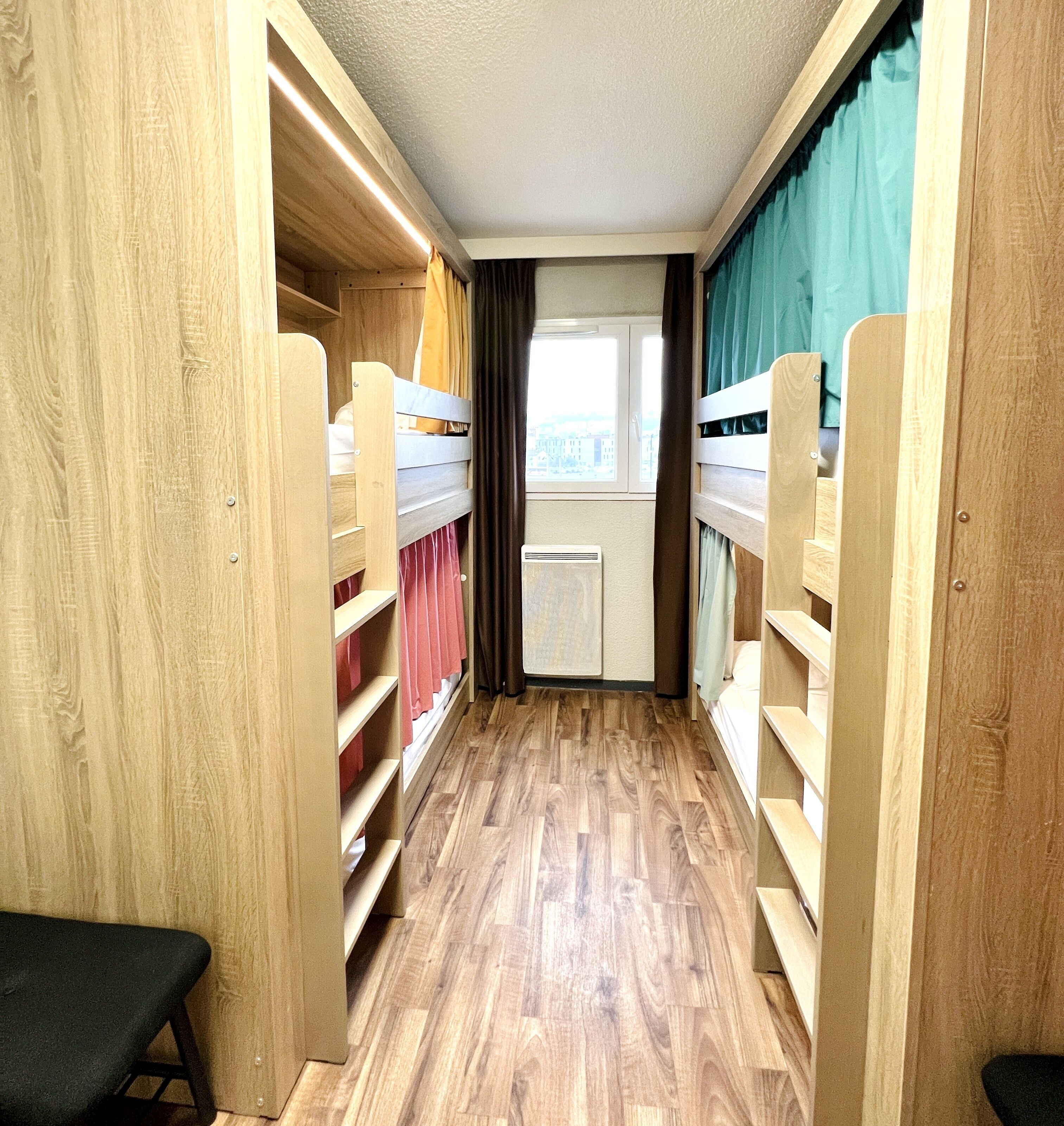 A narrow hallway with wooden bunk beds on either side, a bench at the end, and a window at the far end.