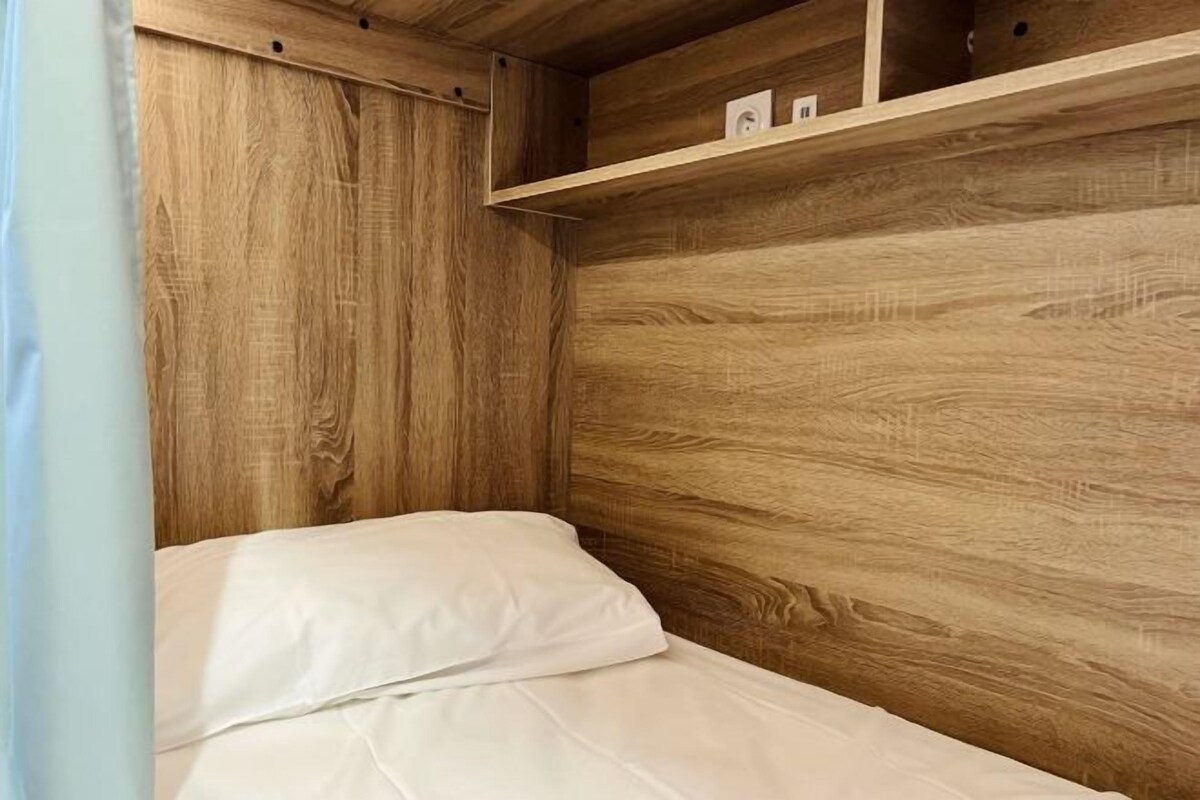A wooden bunk bed with white bedding, a shelf above the bed, and a small white object on the shelf.