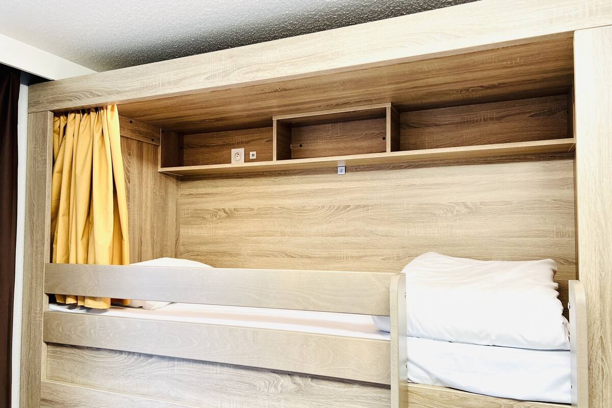 Een stapelbed met houten hoofdbord en planken, een geel gordijn en een wit kussen.