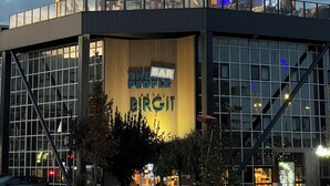 Een modern gebouw met een grote glazen gevel, een terras op het dak met lichtslingers, en een bord waarop staat 
