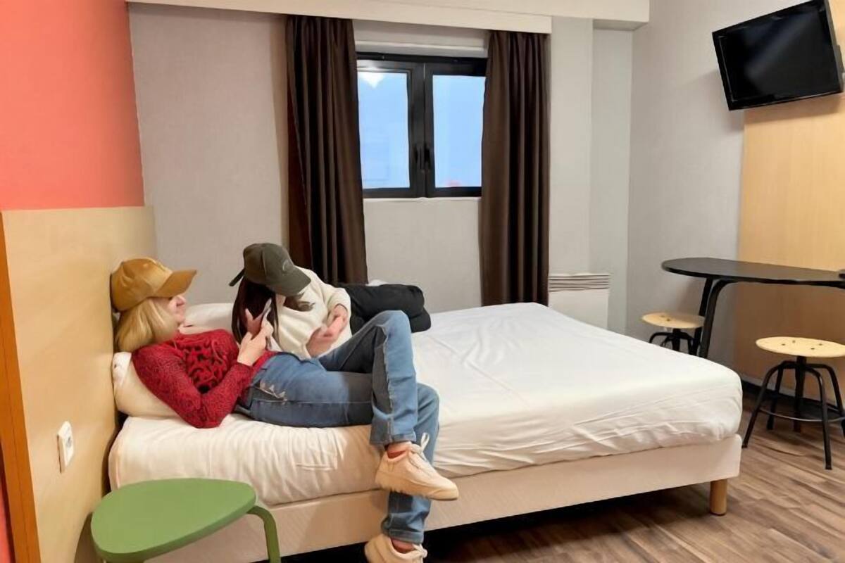 Een hotelkamer met een bed, een kleintje tafeltje, een aan de muur bevestigde televisie en een raam met gordijnen.