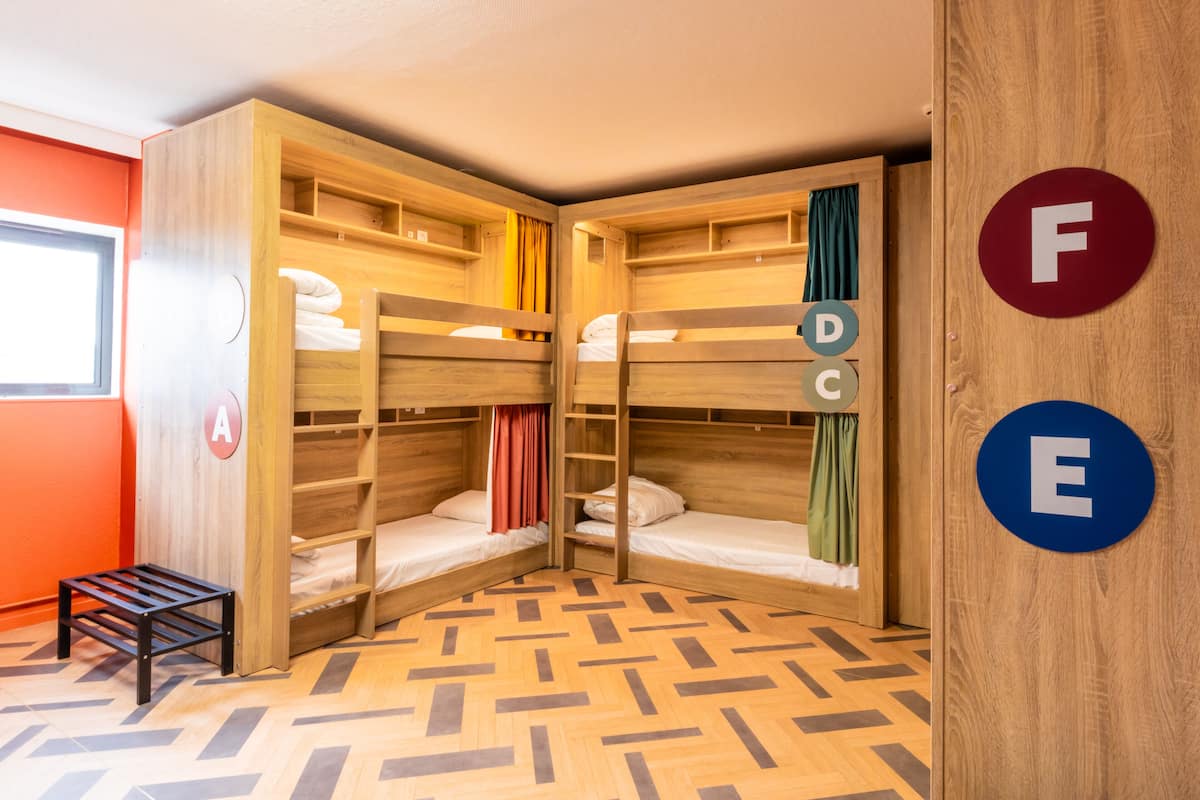 Een kamer met stapelbedden, een klein krukje en een raam. Op de muren staan labels met de letters A, D, C, F en E.