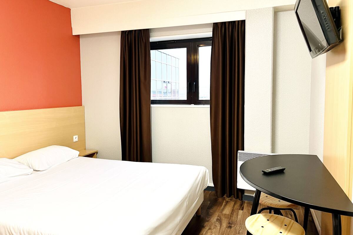 Hotelkamer met een bed, een klein tafeltje, een stoel, een aan de muur bevestigde televisie en een raam met gordijnen.