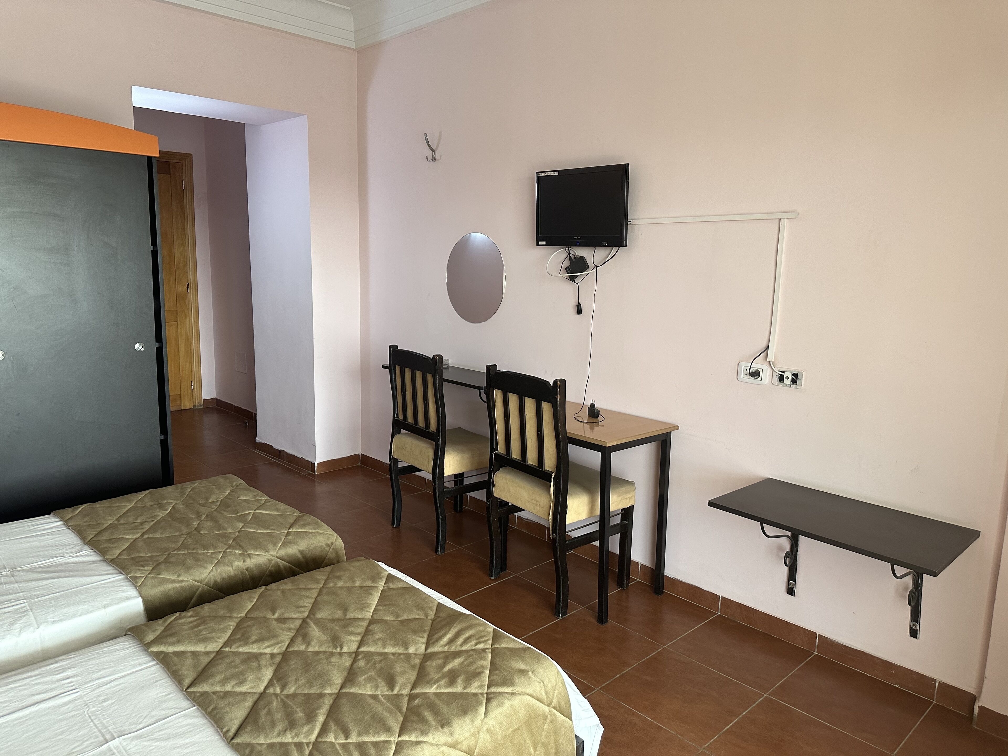 Una habitación de hotel con dos camas, un escritorio con sillas, un televisor y un estante pequeño.