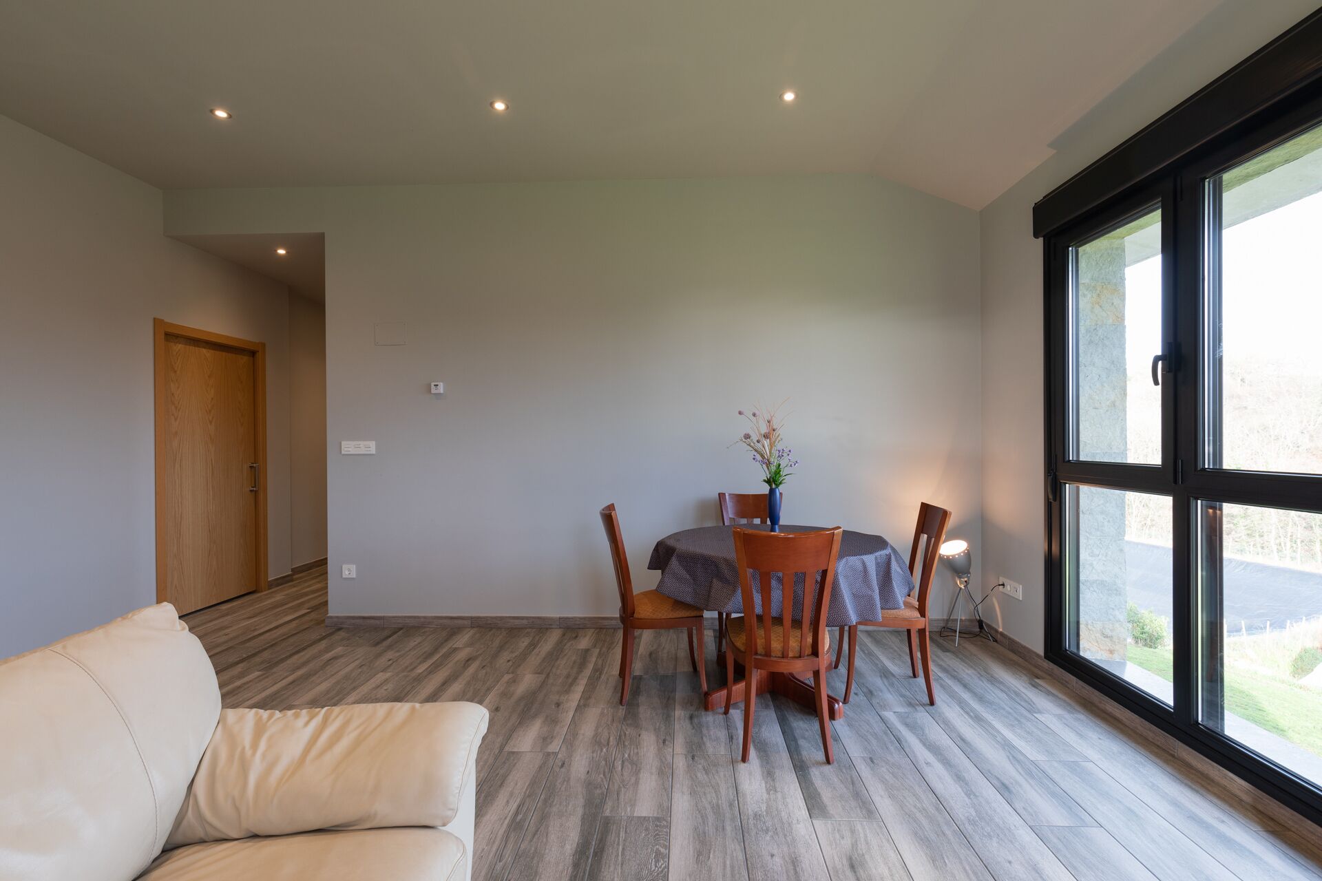 Un comedor con una mesa redonda, cuatro sillas y un florero. Se observa una puerta de madera y un ventanal grande con vistas al exterior.