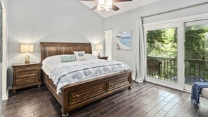 Un dormitorio con una cama grande, cabecera de madera, dos mesitas de noche con lámparas, ventilador de techo y un ventanal con vistas a zonas verdes.