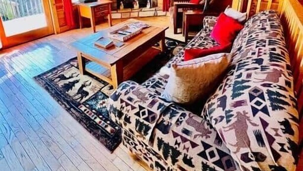 L’intérieur d’une cabane en rondins, avec une cheminée en pierre, un téléviseur fixé au-dessus, un canapé agrémenté de coussins à motifs et un plancher en bois.