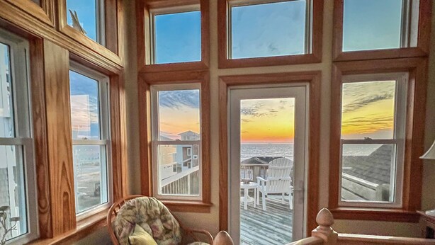 Una habitación con ventanas amplias, una silla de madera y vista a un atardecer sobre un muelle.